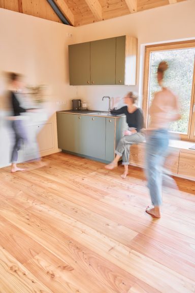 Drei Frauen bewegen sich lebhaft in einem hellen Raum mit Holzfußboden und moderner Küche.
