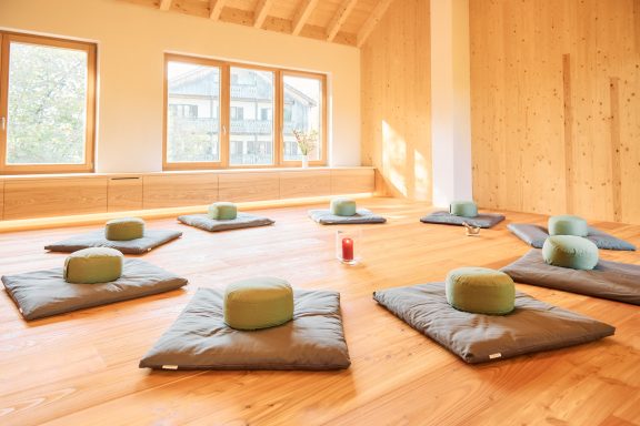 Meditationsraum mit Mats und Kissen auf Holzboden, helle Fenster im Hintergrund.